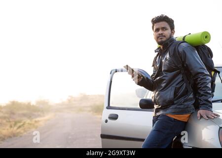 Young traveler using mobile while waiting outside the car with backpack - Cocept of car failure or broken during travel on raodside. Stock Photo
