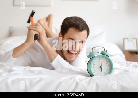 Angry young man with alarm clock and hammer in bedroom Stock Photo