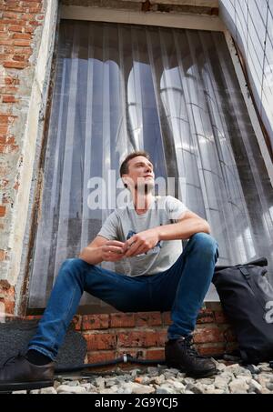 Below view of smiling male sitting on stairs of abandoned house. Young man is resting on stairs outdoor. Concept of leisure. Stock Photo