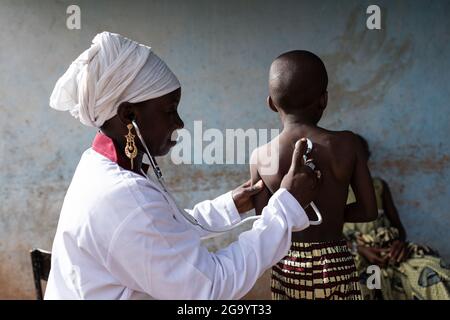 In this image a young smiling female pediatrician is pressing a stethoscope on a small African boy's back to listen to his breathing sounds during a p Stock Photo