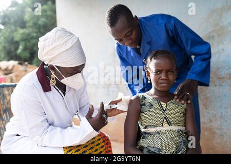 In this image a frightened little black African girl being vaccinated by a female nurse with face mask is comforted by a man in a blue coat bending ov Stock Photo