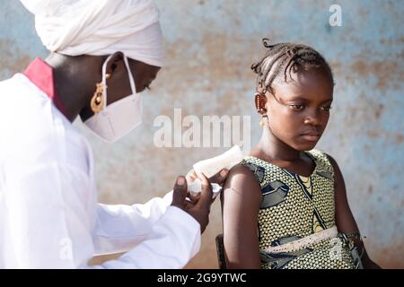 In this image, a young black nurse in a white coat wearing a protective mask is injecting an intramuscular vaccine into the arm of a small serious sch Stock Photo