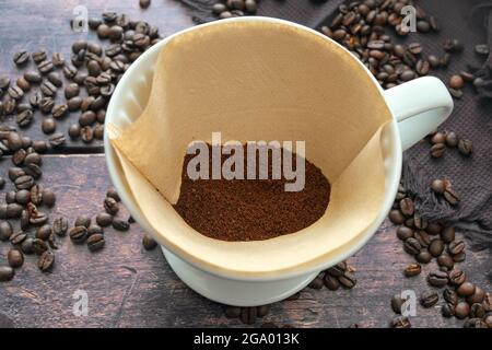 Ground coffee in a paper filter bag and in a porcelain holder for an aromatic drip brewed hot drink, rustic wooden table with some beans, selected foc