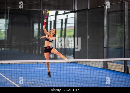 Woman playing padel in a blue grass padel court indoor - Young sporty woman padel player hitting ball with a racket Stock Photo