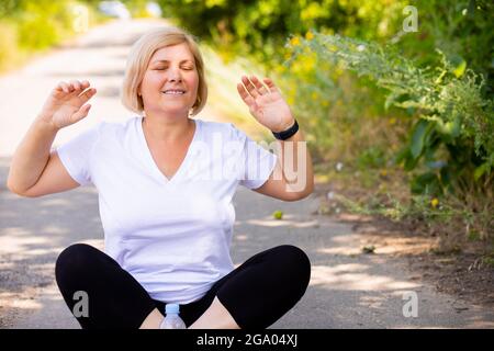 A woman in white t-shirt and black leggings sitting on yoga position outdoors doing meditation with hands keeping up. Stock Photo