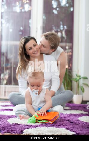 A beautiful little baby sitting on the floor and playing with a toy. Happy parents in the background. Father kissing a mother. Stock Photo