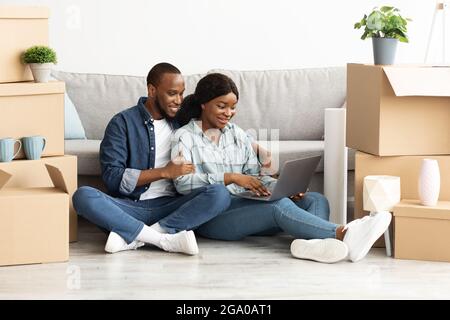 Smiling Black Sposes Sitting With Laptop Among Cardboard Boxes In New House Stock Photo