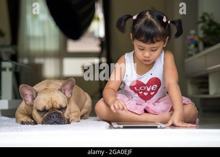 Adorable Southeast-Asian little girl sitting with dog looking at tablet Stock Photo