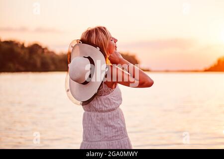 Portrait of a young woman on the background of a sunset. A beautiful happy blonde in a lace summer dress and a straw hat decorated with a ribbon and Stock Photo