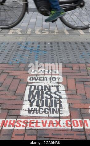 London, UK, 28 July 2021: In Windrush Square, Brixton, a painted sign on the pavement at a pedestrian crossing reminds anyone over the age of 18 that they are eligible for a coronavirus vaccination. Vaccine take-up is lower in big cities than in the rest of the country. Anna Watson/Alamy Live News Stock Photo