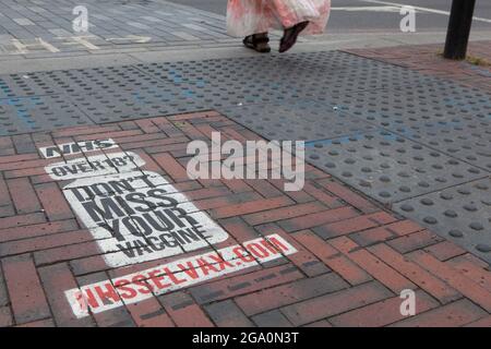 London, UK, 28 July 2021: In Windrush Square, Brixton, a painted sign on the pavement at a pedestrian crossing reminds anyone over the age of 18 that they are eligible for a coronavirus vaccination. Vaccine take-up is lower in big cities than in the rest of the country. Anna Watson/Alamy Live News Stock Photo