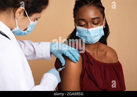 Nurse in workwear making vaccination for black lady Stock Photo