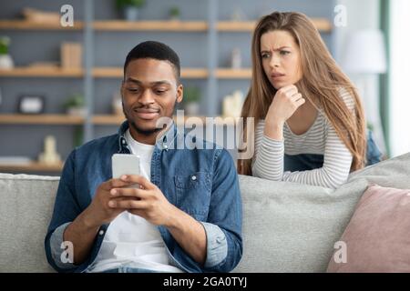 Mistrust. Attentive Lady Looking At Black Boyfriend While He Texting On Smarthone Stock Photo
