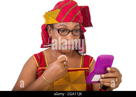 beauty, makeup and elderly concept in red african outfit - senior woman applying powder to face looking in small mirror on white background Stock Photo