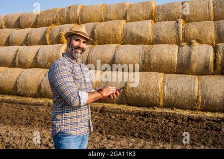 Happy farmer is standing beside bales of hay. He is examining straw after successful harvesting. Stock Photo