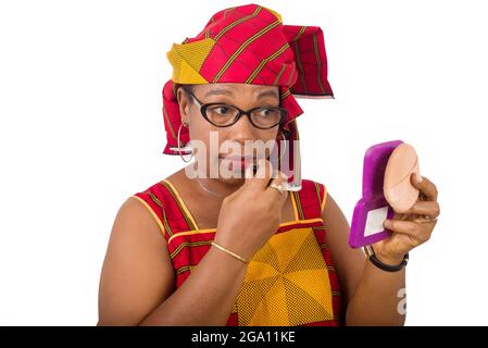 beauty, makeup and elderly people concept in red african outfit - smiling senior woman applying lipstick on her lips looking in small mirror on white Stock Photo