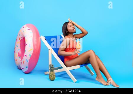 Full length of young black lady suffering from summer heat, holding tropical cocktail while sitting in lounge chair Stock Photo