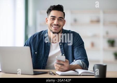 Smiling arab guy using smartphone while sitting at workdesk Stock Photo