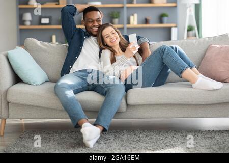 Cheerful multicultural spouses relaxing on couch with smartphone together Stock Photo