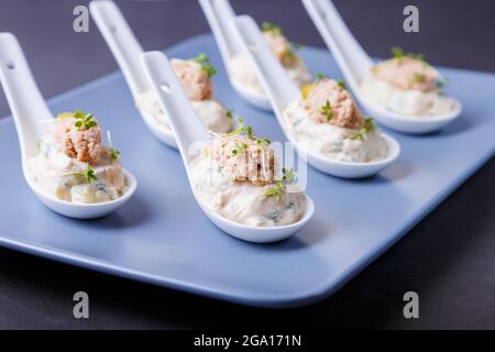 Cold appetizer made from cod liver, cod caviar, olives, cucumber and microgreens on serving spoons. Traditional cold dish. Close-up, blue plate, black Stock Photo