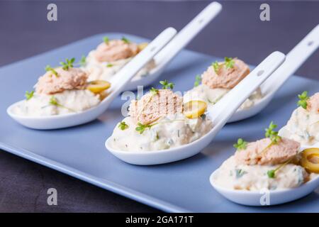 Cold appetizer made from cod liver, cod caviar, olives, cucumber and microgreens on serving spoons. Traditional cold dish. Close-up, blue plate, black Stock Photo