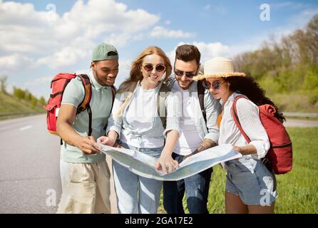 Group of best friends with backpacks checking map during road trip, hitchhiking on highway Stock Photo