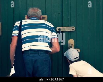 Senior and child are looking through the mail slot and keyhole, view from the back. Grandfather and grandson see something interesting Stock Photo