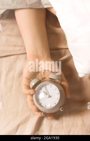 Hands of a young man from under the blankets hold an alarm clock in gray.  Stock Photo