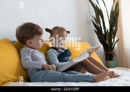 Two preshool, toddler children read large interesting book of fairy tales on yellow bed. Siblings little twins reader have fun, happy kid on Stock Photo