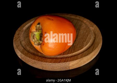 One juicy organic persimmon on a round wooden tray, close-up, against a black background.