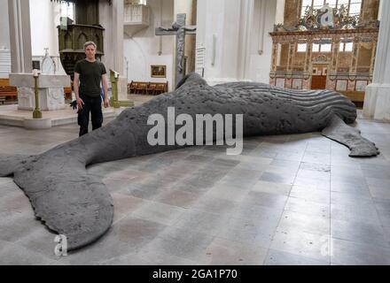 Greifswald, Germany. 28th July, 2021. Gil Shachar, artist from Isarel, stands next to the whale sculpture of a humpback whale in St. Nicholas Cathedral. The 14-metre-long whale sculpture is a cast of an animal washed up in South Africa in 2018. The art project in Greifswald is intended to make people think. On it, for example, bite marks from sharks or also cuts from ship's propellers can be seen. The sculpture has already been exhibited in Berlin and Bochum. Credit: Stefan Sauer/dpa/Alamy Live News Stock Photo