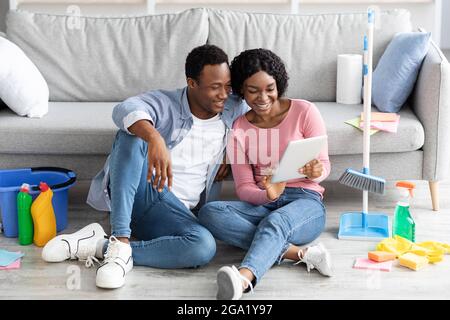 Smiling black couple with cleaning tools resting, using digital tablet Stock Photo