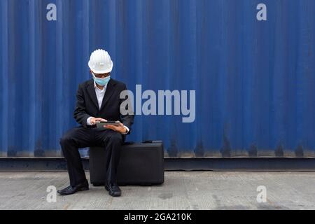 asian manager businessman wearing face mask to protect form covid or coronavirus sitting on bag and working seriously on tablet with worrying business Stock Photo