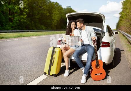 Young couple traveling by auto, sitting in car trunk with suitcase and guitar, holding map, checking destination Stock Photo