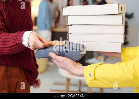 Young smiling blond female in smart casualwear holding credit card over screen of payment machine in hand of male courier in yellow uniform Stock Photo