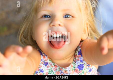 Little blonde preschool girl reaches up with both hands, looks at a camera. A blue-eyed cute female child 2-3 years old smiles with open mouth and ton