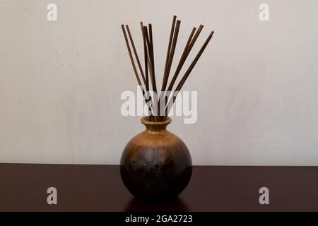A reed diffuser made up of a brown jar with the oils and many bamboo sticks. The bottle sits on a wooden table against a white wall. Stock Photo