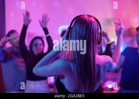 Back view of young woman with long blond hair touching headphones while standing in front of crowd of excited friends dancing at party Stock Photo