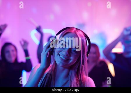 Young cheerful woman with long blond hair snapping fingers while enjoying music in headphones in front of dancing crowd at party Stock Photo