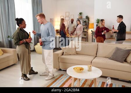 Happy young intercultural couple with bottles of beer looking at you while enjoying home party against their friends chatting and having drinks Stock Photo