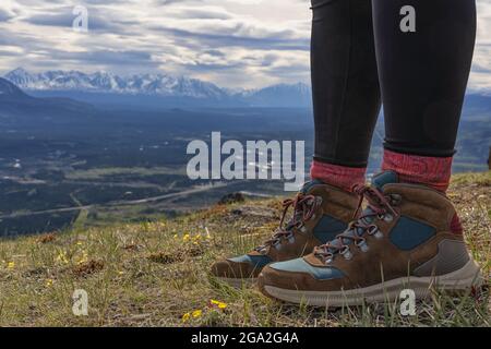 Close-up of woman's feet wearing hiking boots, standing on a mountaintop with a vista of the mountain ranges and valley below in the distance on a ... Stock Photo