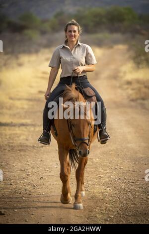 Woman riding horse (Equus ferus caballus) on a dirt road through the bush on the savanna looking at the camera and smiling at the Gabus Game Ranch Stock Photo