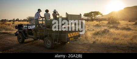 View taken from behind of a safari guide and women travelers standing in a parked jeep talking and looking out onto the savanna with binoculars on ... Stock Photo