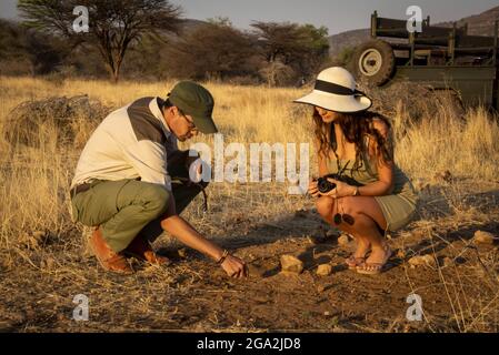 Safari guide with woman traveler wearing a straw hat and holding a camera crouching down on the ground talking on the savanna at the Gabus Game Ran... Stock Photo