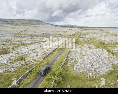 Aerial view of a car driving down a road through the barren, lunar landscape of limestone found in the Burren National Park with vegetation growing... Stock Photo