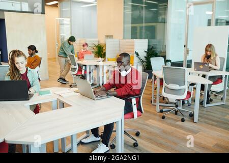 Young serious office managers or designers of various ethnicities sitting by desks and using laptops during individual work in open space Stock Photo