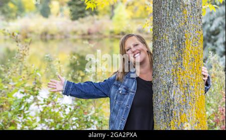 A middle aged woman holding a tree and reaching her hand out while enjoying herself on a warm fall evening in a city park; Edmonton, Alberta, Canada Stock Photo