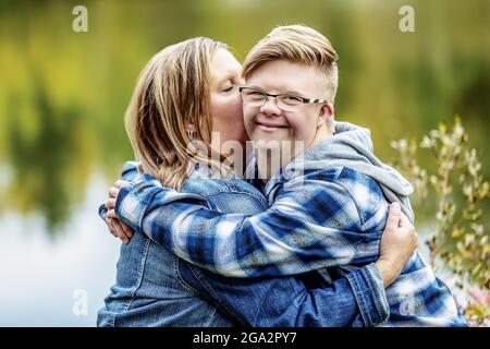 A young man with Down Syndrome being hugged by his mother while enjoying each other's company in a city park on a warm fall evening Stock Photo