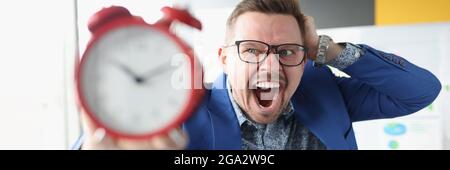 Businessman in glasses holding red alarm clock in his hands and shouting Stock Photo
