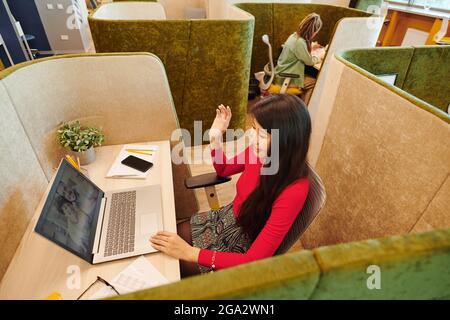 Asian young businesswoman in smart casualwear and protective mask waving hand while looking at happy couple on laptop display Stock Photo
